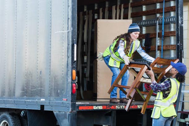 Two multi-ethnic female workers in their 30s and 40s at the back of a truck, loading or unloading furniture. The women are wearing reflective vests and jeans. They are moving merchandise for a furniture store.