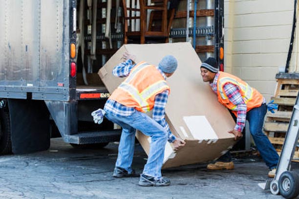 Two multi-ethnic mature workers in their 40s at the back of a truck, loading or unloading a large cardboard box. The men are wearing plaid shirts, reflective vests and jeans. They are moving merchandise for a furniture store.